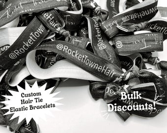 May include: A pile of black elastic hair ties with white text that reads "@RocketTown" and "@RocketTowneHair". The text "Custom Hair Tie Elastic Bracelets" and "Bulk Discounts!" are also visible in the image.
