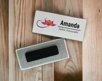 以下が含まれることがあります： 紫色の花のロゴと「Amanda Sales Associate」というテキストが書かれた白い長方形のネームタグ。ネームタグは木製の表面に置かれています。銀色の背景に黒い警告ラベルが貼られており、「CAUTION MAGNETIC DEVICE DO NOT USE WITH PACEMAKER」と書いてあります。