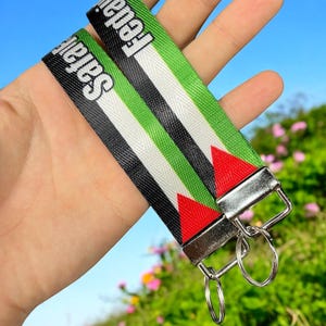 May include: A black and green key chain with the words "Palestine" and "Free Palestine" printed on it. The key chain has a silver metal clasp and two silver rings.