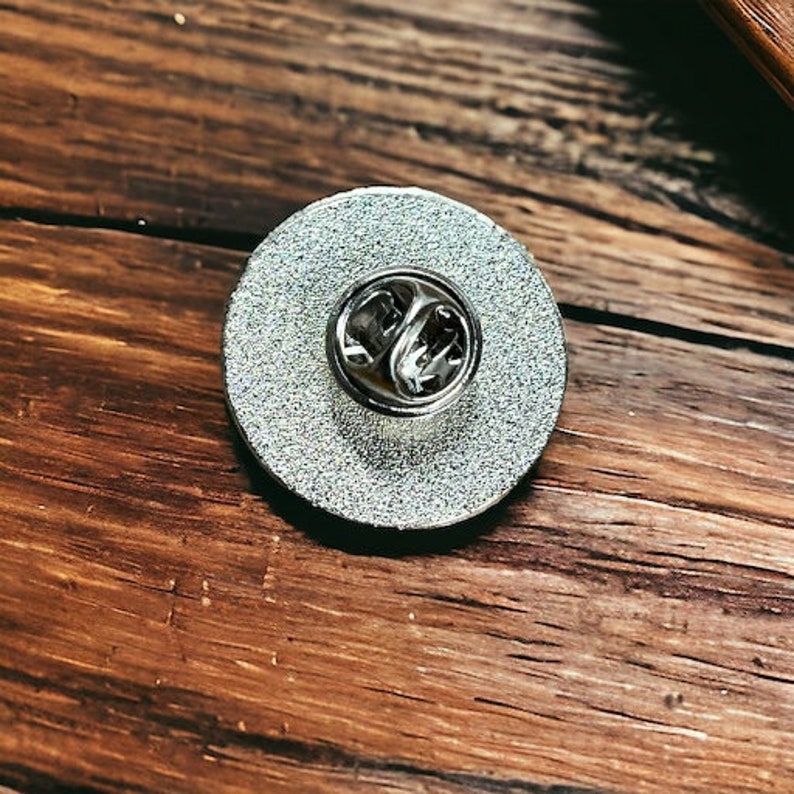 May include: A circular silver-toned metal pin badge featuring a textured, glittery finish on the back. The center of the round disc displays a standard metal butterfly clutch fastener mechanism, designed to secure the pin to fabric or garments.