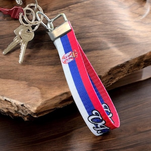 May include: A white, blue and red keychain with the Serbian flag and the word "Serbia" printed on it. The keychain is attached to a silver key ring.