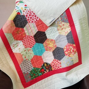 May include: A white baby quilt with a red border and a patchwork design of hexagons in various colors and patterns.