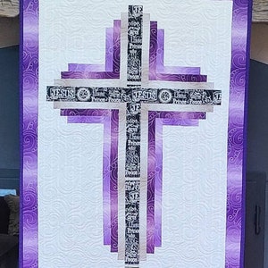 May include: Purple quilted wall hanging with a white cross in the center. The cross is made of multiple layers of fabric with the words "JESUS", "Prince", "Peace", "King", "Lord", "God", and "TE" printed on it.