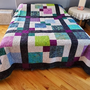 May include: A colorful patchwork quilt with squares of green, purple, blue, and white fabric. The quilt is laid out on a bed with a wooden floor.