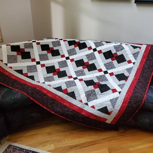 May include: A handmade quilt with a geometric pattern in black, white, gray, and red. The quilt features a repeating square design with a red border and a dark patterned edge. The quilt is draped over a dark sofa.