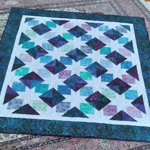 May include: A handmade quilt with a white center featuring a geometric pattern of diamonds, squares, and triangles in shades of blue, purple, and teal. The quilt has a dark teal border with a patterned design.
