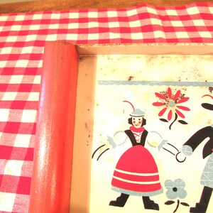 Puede incluir: Una bandeja de metal vintage con un dise&ntilde;o colorido que presenta dos figuras con ropa tradicional, una flor y una flor azul y blanca. La bandeja tiene un asa de madera roja.