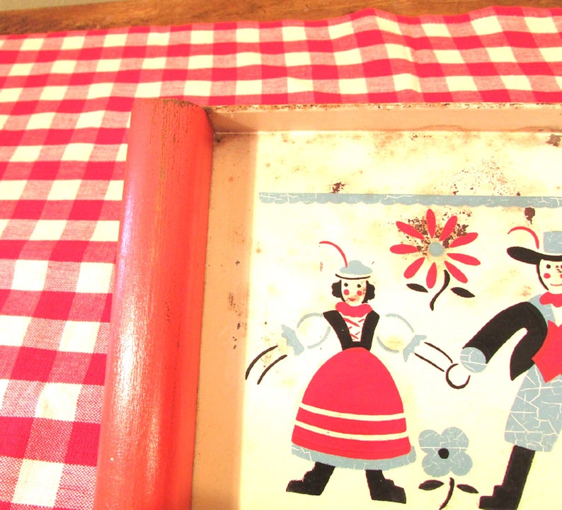 Puede incluir: Una bandeja de metal vintage con un dise&ntilde;o colorido que presenta dos figuras con ropa tradicional, una flor y una flor azul y blanca. La bandeja tiene un asa de madera roja.