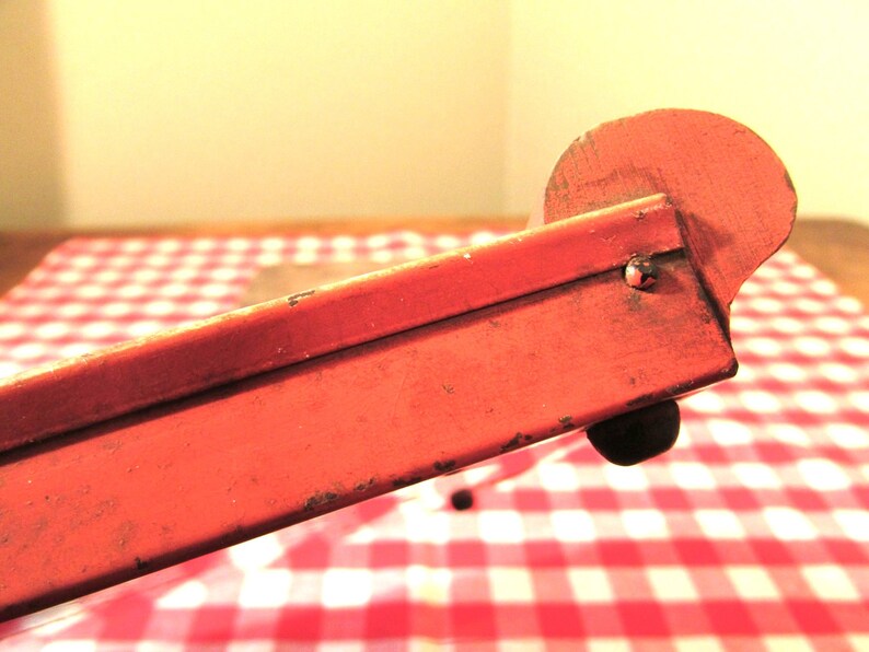 Puede incluir: Un juguete de metal rojo con una rueda en un extremo. El juguete est&aacute; sobre un mantel a cuadros rojo y blanco.