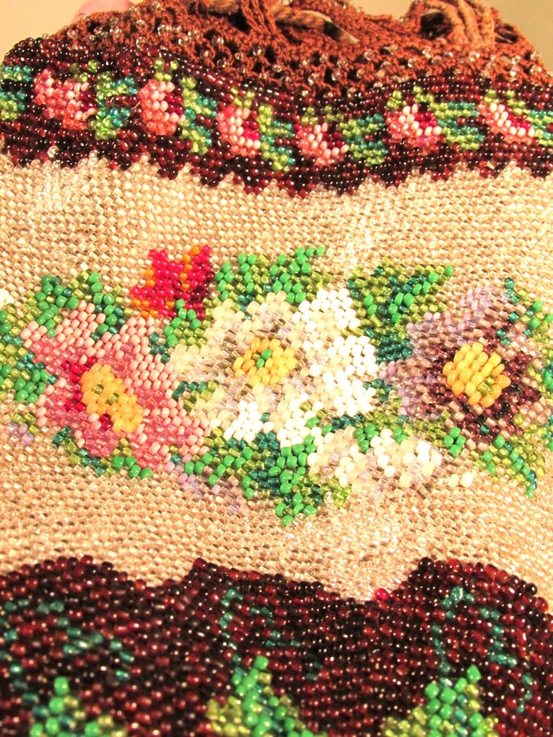 May include: A close-up of a beaded purse with a floral design. The purse is made of brown, green, white, pink, and yellow beads. The beads are arranged in a pattern of flowers and leaves.