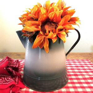 May include: A vintage-style, two-tone gray and white enamelware pitcher with a black handle, filled with vibrant orange sunflowers. The pitcher sits on a red and white checkered tablecloth, with a red bandana in the foreground.