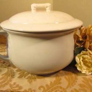 May include: White ceramic chamber pot with a lid and a handle. The lid has a small handle on top. The pot is sitting on a patterned fabric surface.