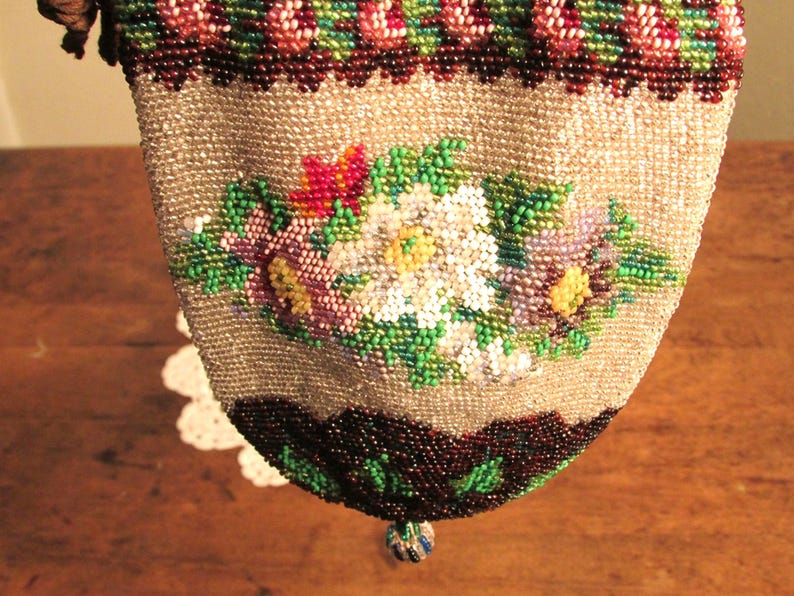 May include: A beaded purse with a floral design in shades of pink, white, green, and purple. The purse is made of small, iridescent beads and has a drawstring closure.