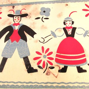 Puede incluir: Una fiambrera vintage de hojalata con un dise&ntilde;o pintado de dos figuras en trajes tradicionales folcl&oacute;ricos bailando. Las figuras est&aacute;n rodeadas de flores estilizadas en rojo, azul y blanco.