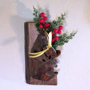 May include: A rustic wall hanging featuring a piece of textured wood with a decorative burl. It is adorned with faux evergreen sprigs, bright red berries, and a small pine cone, tied with a yellow ribbon. The wood is mounted on a rectangular wooden base.