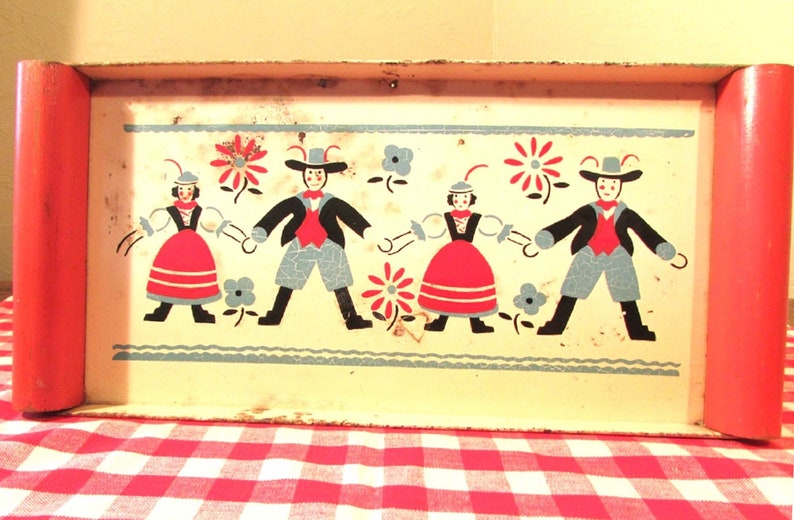 Puede incluir: Una bandeja de metal vintage con borde rojo e interior blanco. La bandeja presenta un dise&ntilde;o colorido de cuatro figuras con ropa tradicional, bailando y tom&aacute;ndose de la mano. Las figuras est&aacute;n rodeadas de flores y hojas.
