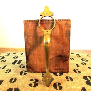 May include: A vintage brass handle with a decorative design, mounted on a wooden box. The handle has a circular grip and a diamond-shaped base. The box is made of dark wood and is set on a burlap runner with black numbers.