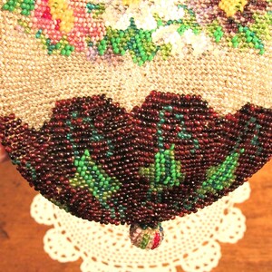 May include: A beaded purse with a floral design in shades of pink, white, and green. The purse has a brown beaded bottom with green accents and a small beaded tassel.
