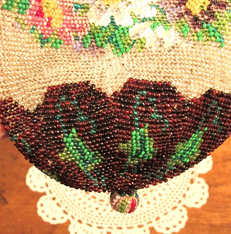 May include: A beaded purse with a floral design in shades of pink, white, and green. The purse has a brown beaded bottom with green accents and a small beaded tassel.