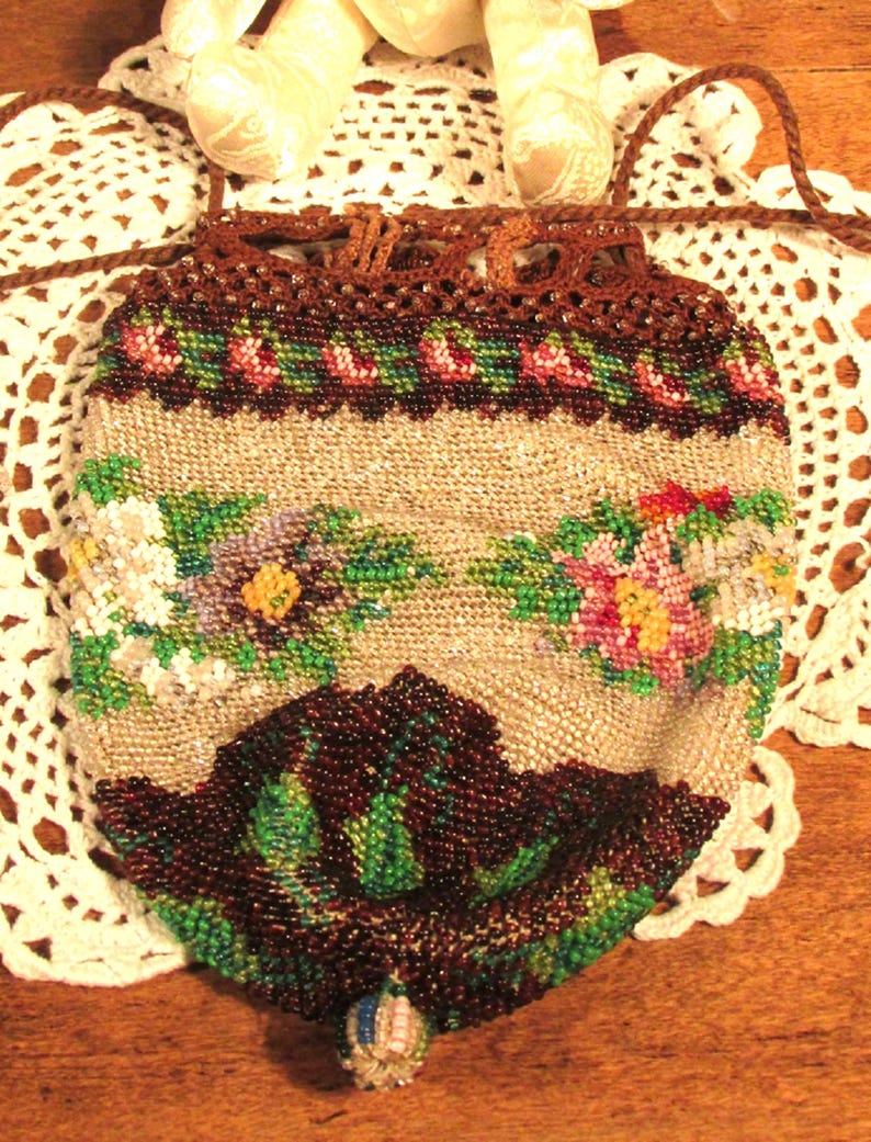 May include: A vintage beaded purse with a floral design in shades of pink, green, and brown. The purse has a drawstring closure and a brown crocheted top.