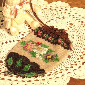 May include: A beaded purse with a floral design in shades of pink, green, and brown. The purse has a brown drawstring closure and is sitting on a white crocheted doily.