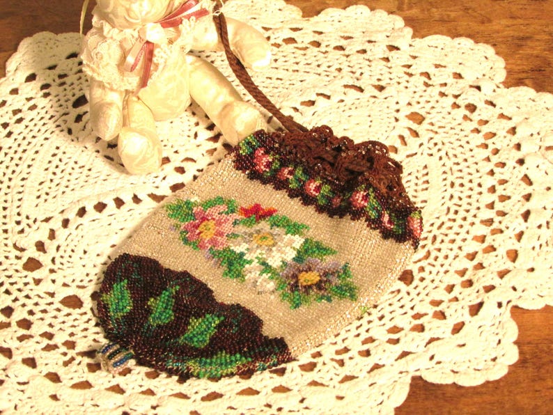 May include: A beaded purse with a floral design in shades of pink, green, and brown. The purse has a brown drawstring closure and is sitting on a white crocheted doily.