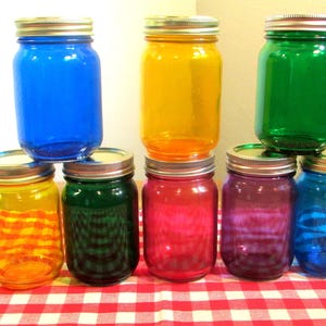 May include: A collection of seven glass mason jars in various colors, including blue, yellow, green, red, purple, and orange. Each jar has a silver lid and is arranged on a red and white checkered tablecloth. The jars are empty and appear to be for decorative purposes.