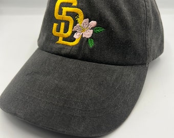 May include: A black baseball cap with a yellow embroidered "SD" and a pink flower.
