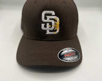 May include: Brown baseball cap with a white embroidered "SD" logo featuring a yellow sun and stars. The cap has a Flexfit label and is size L/XL.