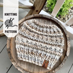 May include: A knitted hat in a beige, white and brown colour scheme. The hat is sitting on a wooden surface. The hat has a small leather patch with a tree logo on it.