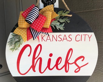 May include: A round wooden door hanger with a black and white striped design. The top half of the door hanger is white with the words "KANSAS CITY" and "Chiefs" in red. The bottom half of the door hanger is black. A yellow and red bow with black and white stripes is attached to the top of the door hanger.