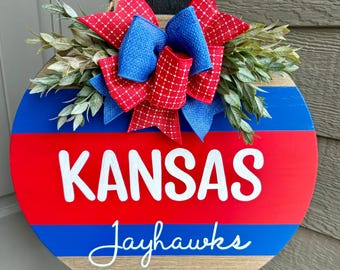 May include: A round wooden door hanger with red, white, and blue stripes. The center of the hanger has the words "KANSAS Jayhawks" in white lettering. A red and blue bow with green foliage is attached to the top of the hanger.
