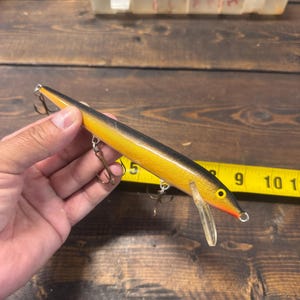 May include: A black and yellow fishing lure with three treble hooks and a clear plastic lip. The lure is held in a hand, with a yellow measuring tape in the background.