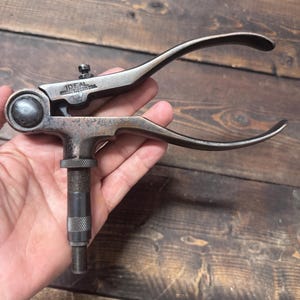 May include: A vintage metal Ideal brand hand tool featuring a curved handle mechanism and a central threaded shaft with knurled adjustment rings. The tool has a dark, weathered patina and includes the stamped text IDEAL SYCAMORE ILL USA.
