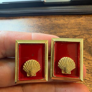 May include: A pair of rectangular cufflinks featuring a polished gold-tone metal frame surrounding a deep red enamel inlay. Centered on the red background of each cufflink is a raised, textured gold-tone seashell motif with detailed ridges.