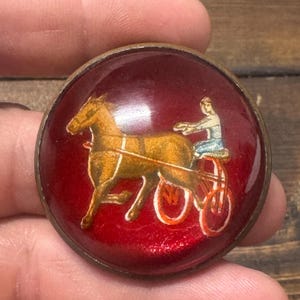 May include: A red glass button with a gold rim features a painted image of a horse pulling a carriage with a person driving. The horse is brown and the carriage has two wheels.
