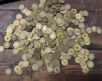 May include: A large pile of vintage brass tokens with a circular design and a central emblem. The tokens are scattered on a dark wooden surface, creating a textured and aged appearance. The tokens appear to be in good condition.