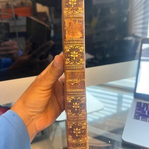 以下が含まれることがあります： 金色の文字と装飾的なパターンが施された、茶色の革張りでアンティークな本の表紙。背表紙には「LONGFELLOW」というタイトルが書かれている。