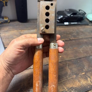 May include: A vintage reloading tool with a metal frame and wooden handles. The tool has a series of circular holes and a screw. The handles are a light brown color and show signs of wear.