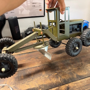 May include: A vintage-style olive green metal toy road grader featuring a central adjustable blade, an open-air operator cabin, and six black rubber tires with deep treads. The construction vehicle has a long frame and a steering wheel inside.