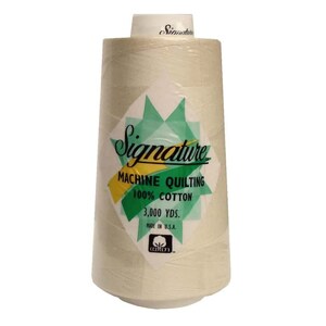 May include: A spool of Signature Machine Quilting 100% cotton thread in a beige color. The spool is labeled with the brand name, "Signature", and the text "Machine Quilting 100% Cotton 3,000 yds. Made in U.S.A."
