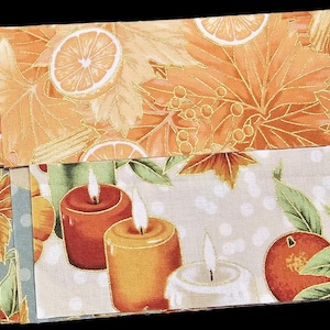 May include: A collection of fabric strips featuring autumn-themed patterns including orange pumpkins, sliced citrus fruits, fall leaves, and lit pillar candles in shades of red, yellow, and white, arranged in a layered, fan-like display.
