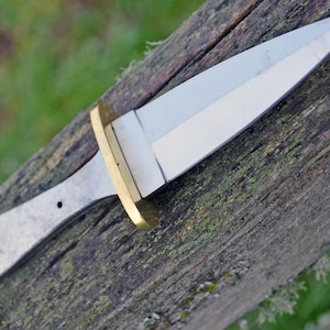 May include: A silver knife with a brass handle. The knife has a sharp point and a straight edge. The knife is lying on a wooden surface.