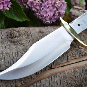 Może przedstawiać: Zbliżenie na srebrne ostrze noża z mosiężnym uchwytem. Nóż leży na kawałku zwietrzałego drewna.