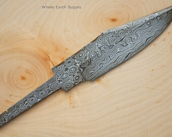 May include: A close-up of a knife blade made of Damascus steel. The blade has a wavy pattern and is about 8 inches long. The blade is lying on a wooden surface.