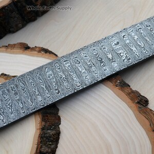 May include: A close-up of a long, rectangular piece of steel with a wavy, patterned surface. The steel is a light gray color with a subtle sheen. The steel is lying on a wooden surface.