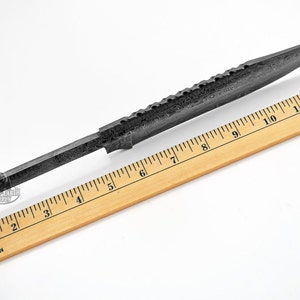 K&ouml;nnte beinhalten: Eine schwarze Stahlmesserklinge mit strukturierter Oberfl&auml;che und einer scharfen Spitze. Die Klinge wird an einem Holzlineal gemessen und ist 30,5 cm lang.