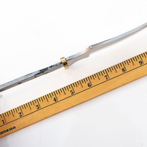 May include: A silver knife blade with a gold ring near the handle. The blade is measured against a wooden ruler with black markings. The ruler is marked with inches from 1 to 10.