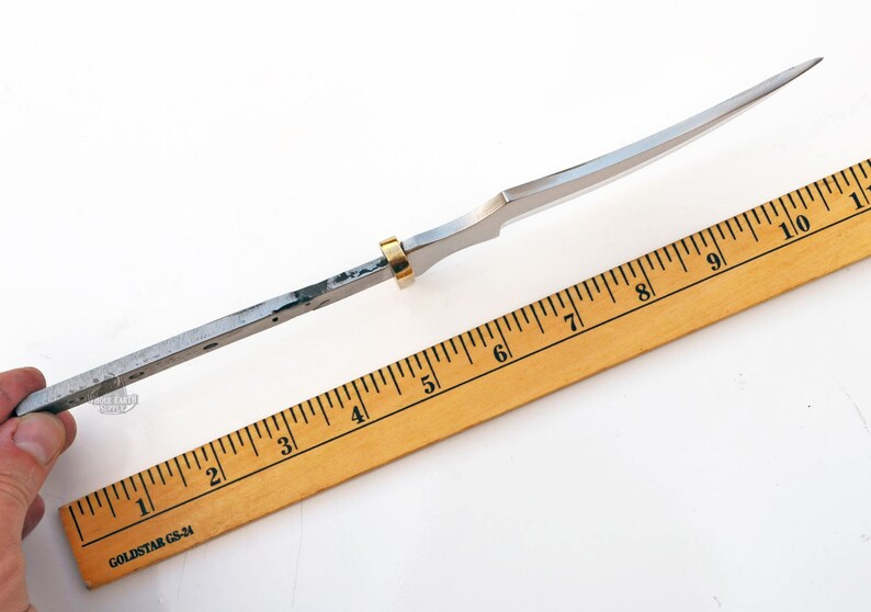 May include: A silver knife blade with a gold ring near the handle. The blade is measured against a wooden ruler with black markings. The ruler is marked with inches from 1 to 10.