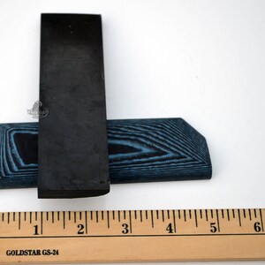 May include: Two black and blue wood blanks for knife making. The blanks are approximately 2 inches long and 1 inch wide. The ruler in the image is marked in inches.